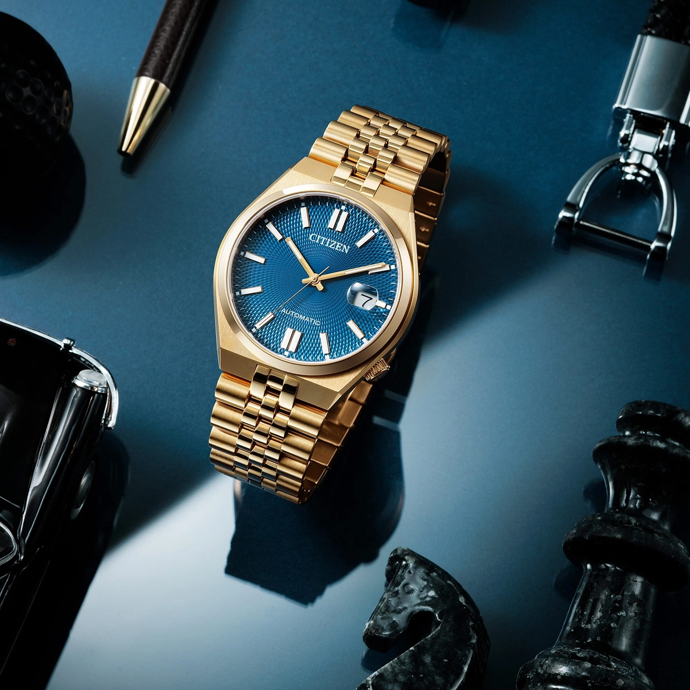 Gouden Citizen horloge met blauwe wijzerplaat, pen, schaakstukken en sleutelhanger op blauw oppervlak.