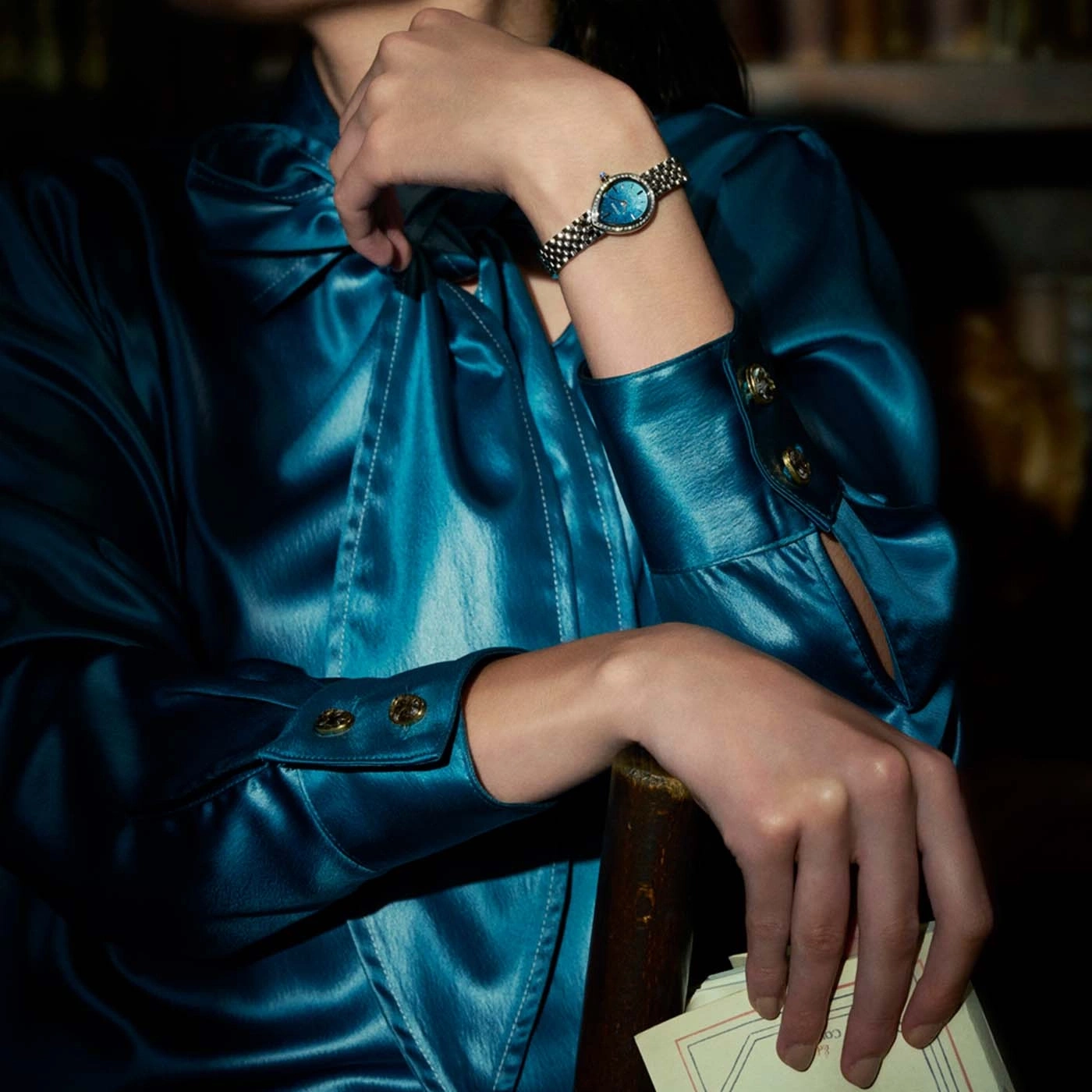 Vrouw in blauwe satijnen jurk met horloge en papieren.