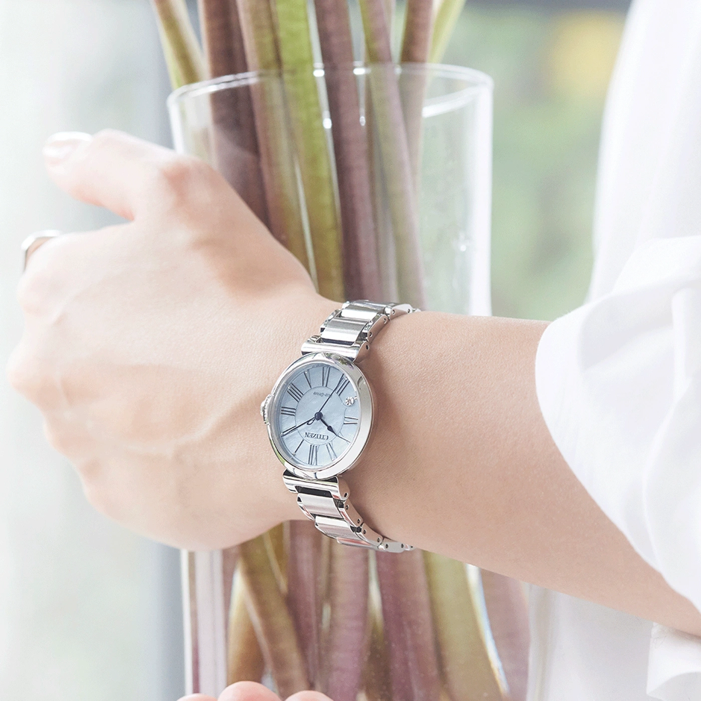 Een pols van een vrouw met een zilveren horloge, die een glazen vaas met planten vasthoudt.