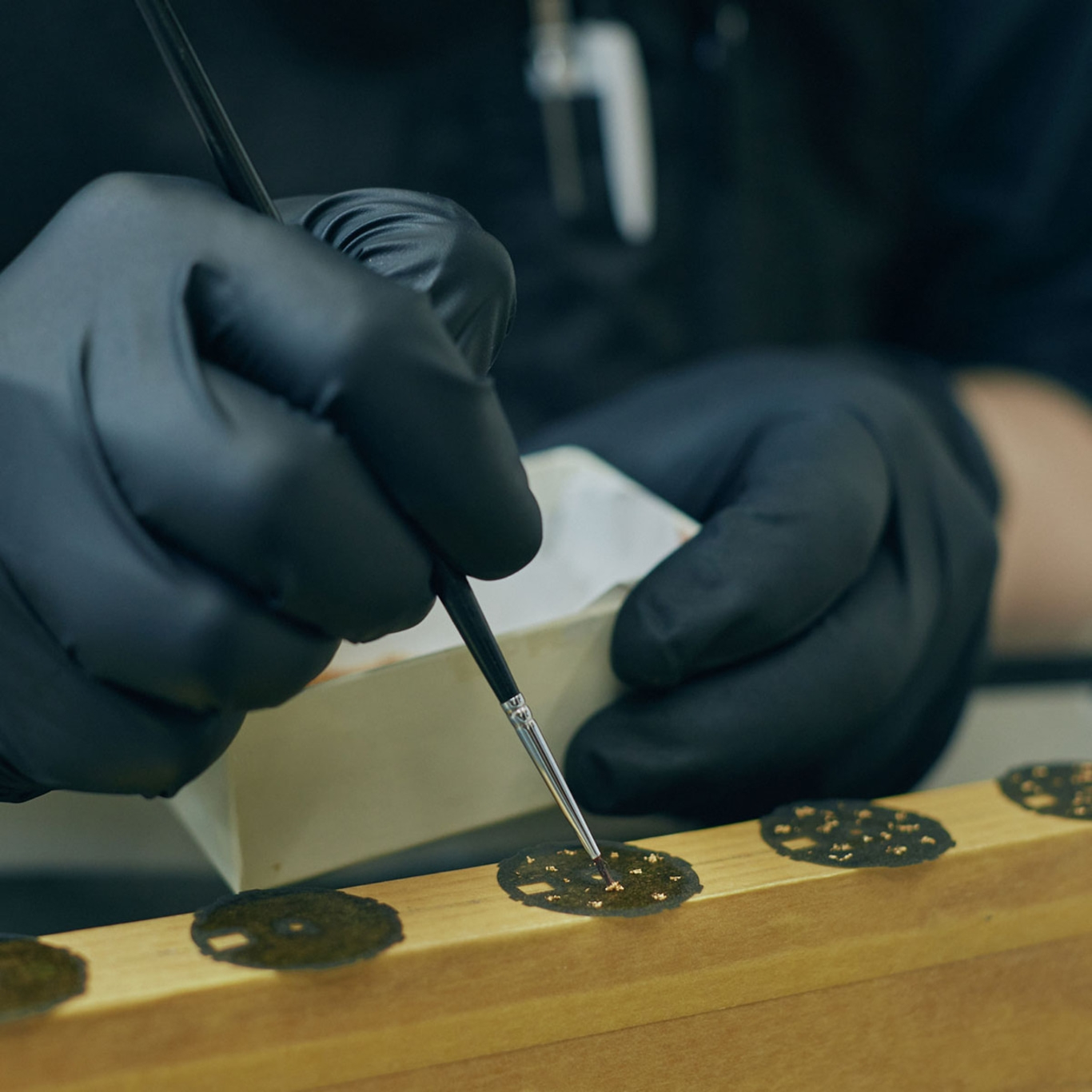 Person in schwarzen Handschuhen bemalt kleine Objekte mit Pinsel auf Holz