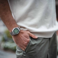 Man draagt zilveren horloge met groene wijzerplaat, hand in zak.