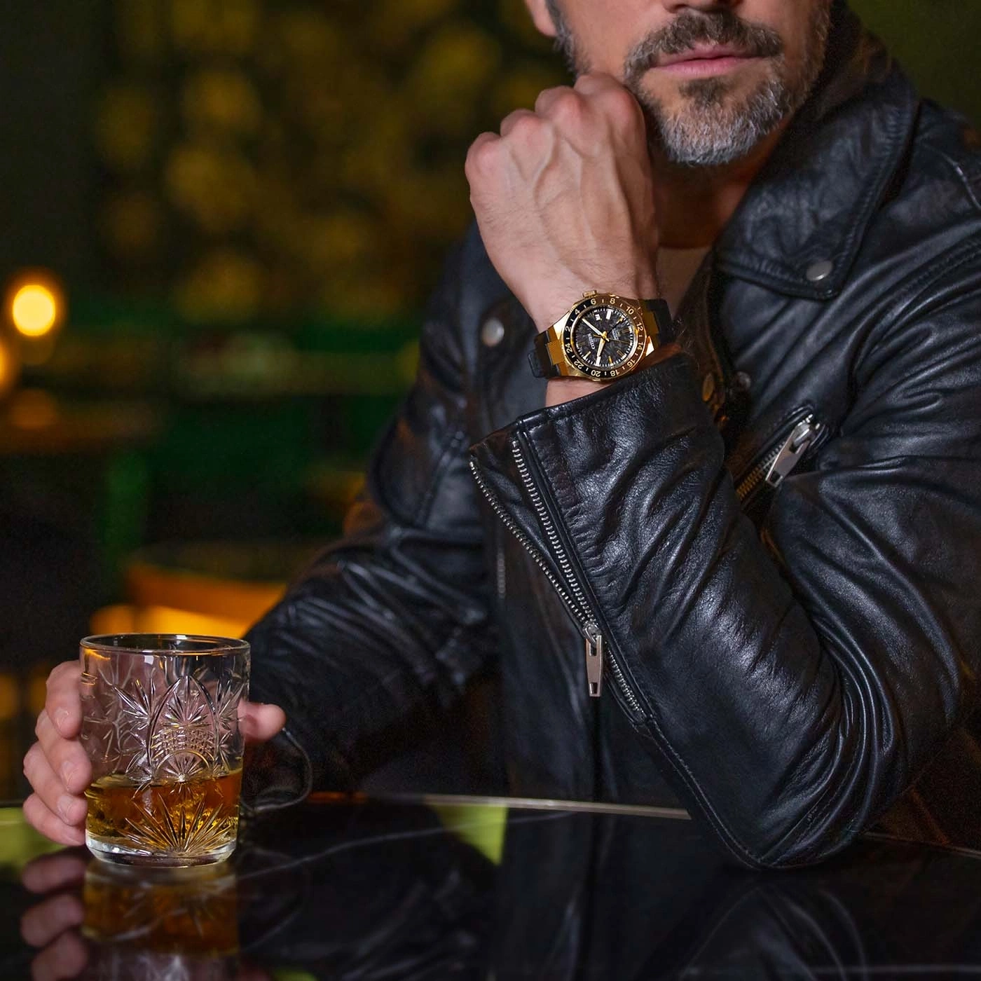 Man in leren jas met horloge houdt whiskeyglas vast.