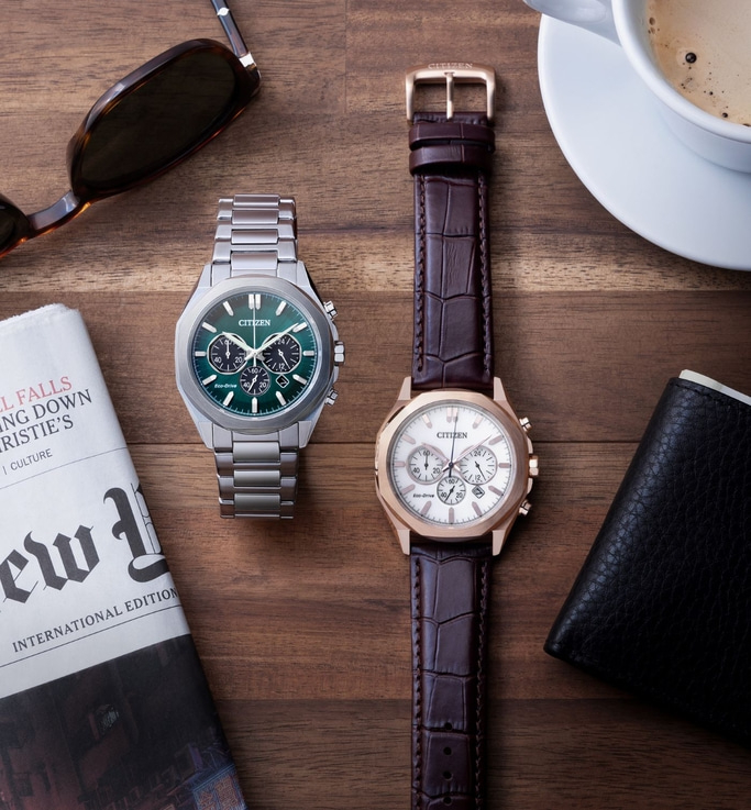 Een stijlvolle flat lay met twee Citizen-horloges, een zonnebril, een krant, een portemonnee en een kop koffie, allemaal gerangschikt op een houten oppervlak.
