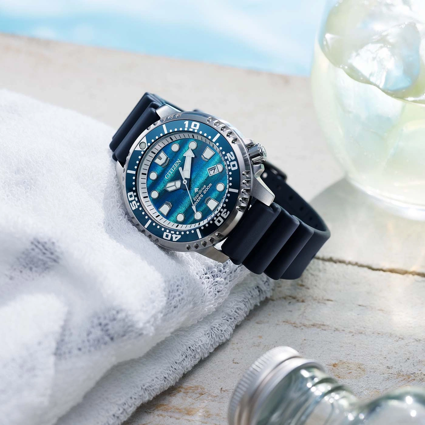 Een blauw Citizen Eco-Drive duikhorloge ligt op een witte handdoek naast een glas met ijsthee.