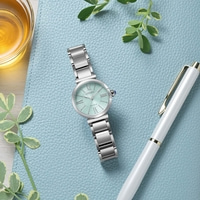Een mintgroen Citizen Eco-Drive horloge met zilveren band ligt op een blauw getextureerd oppervlak naast een witte pen en een glas amberkleurige vloeistof.