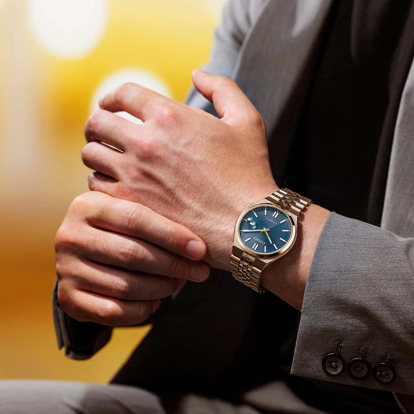 Man draagt gouden horloge met blauwe wijzerplaat en grijs pak.