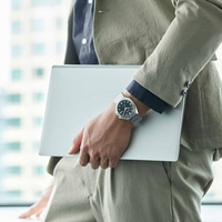 Persoon die een laptop vasthoudt, een horloge en een pak draagt, bij een raam.