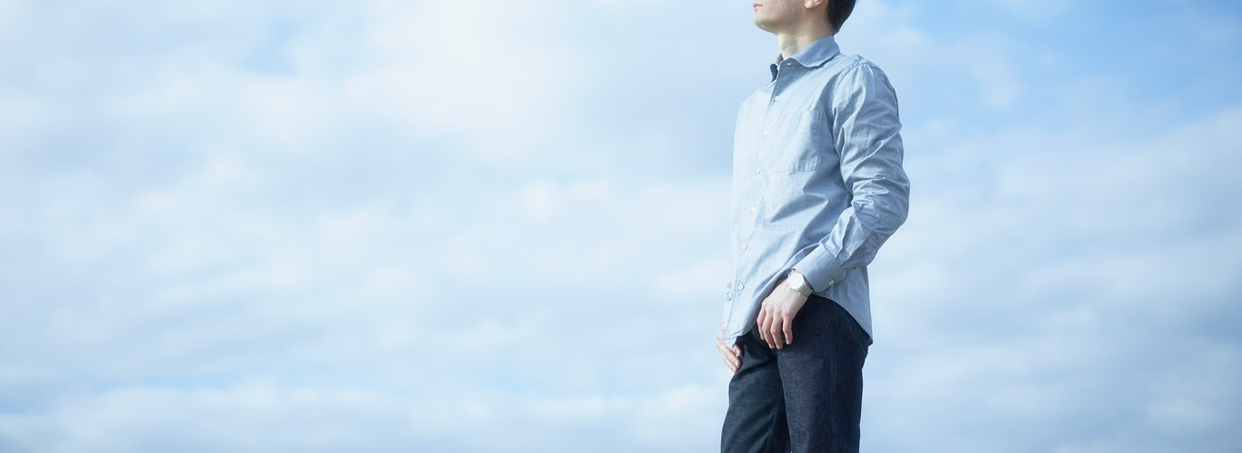 Man in overhemd en jeans staat tegen een bewolkte lucht en kijkt omhoog.