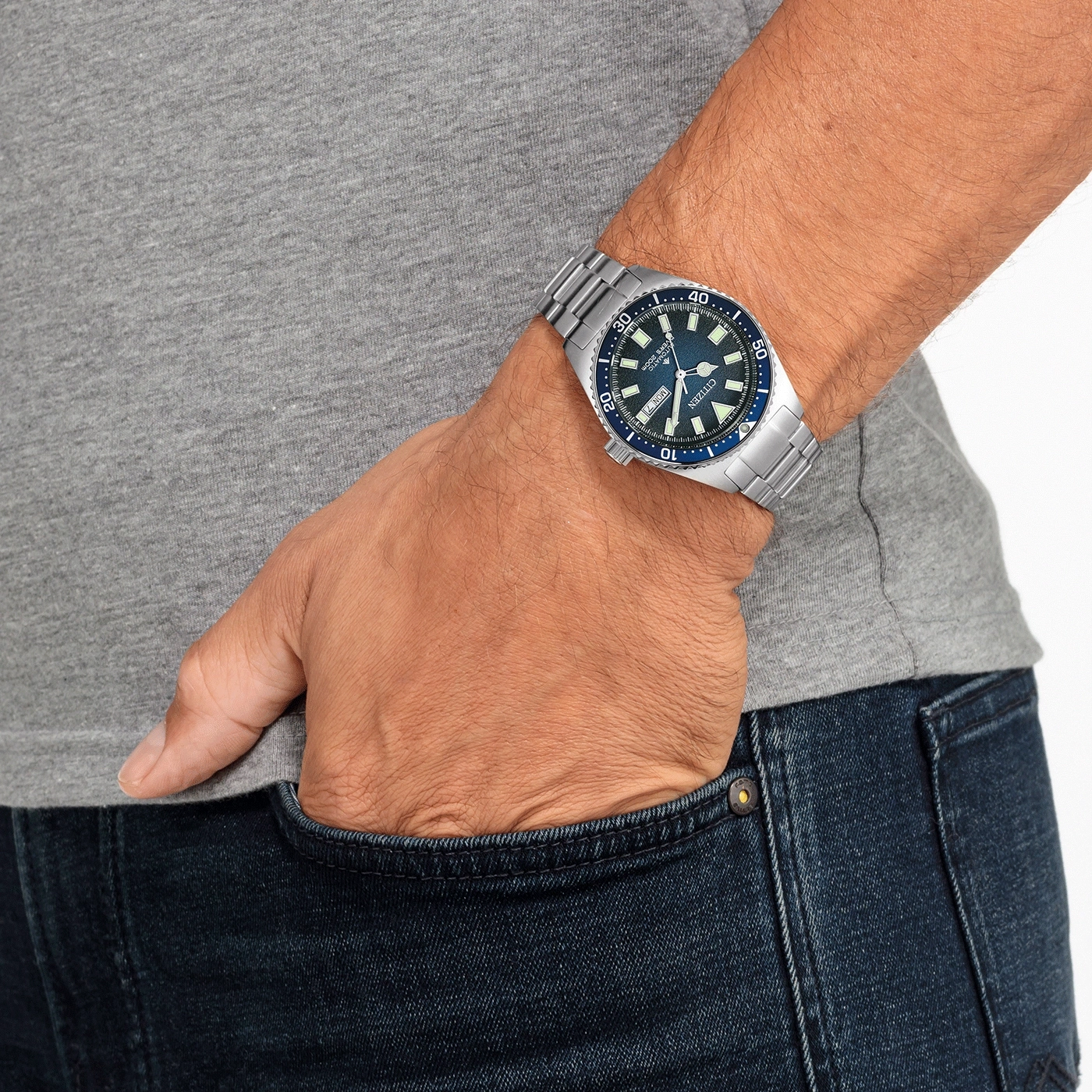 Man met horloge, hand in zak, blauwe jeans en grijs shirt.