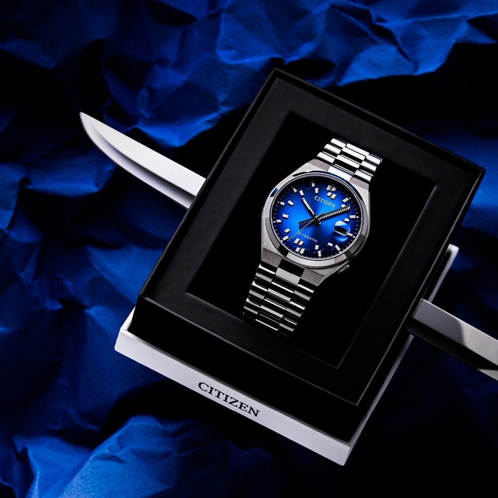 Een blauw Citizen automatisch horloge in een zwarte doos op blauw gekreukt papier.