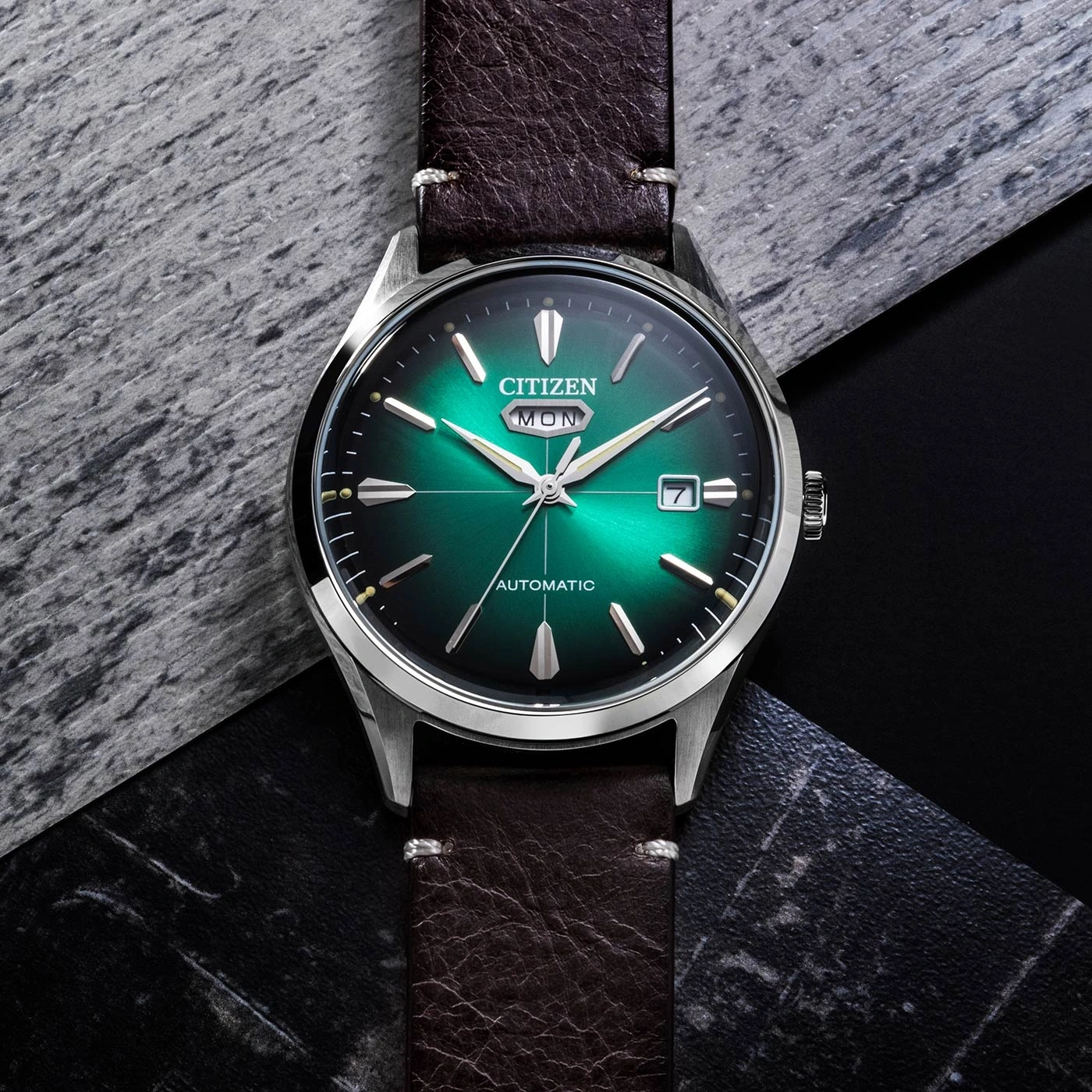 Citizen horloge met groene wijzerplaat en bruine leren band op een getextureerd oppervlak.