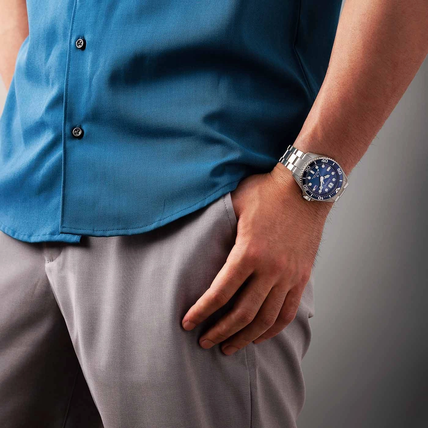 Man met een blauw overhemd en grijze broek met een horloge om zijn pols.