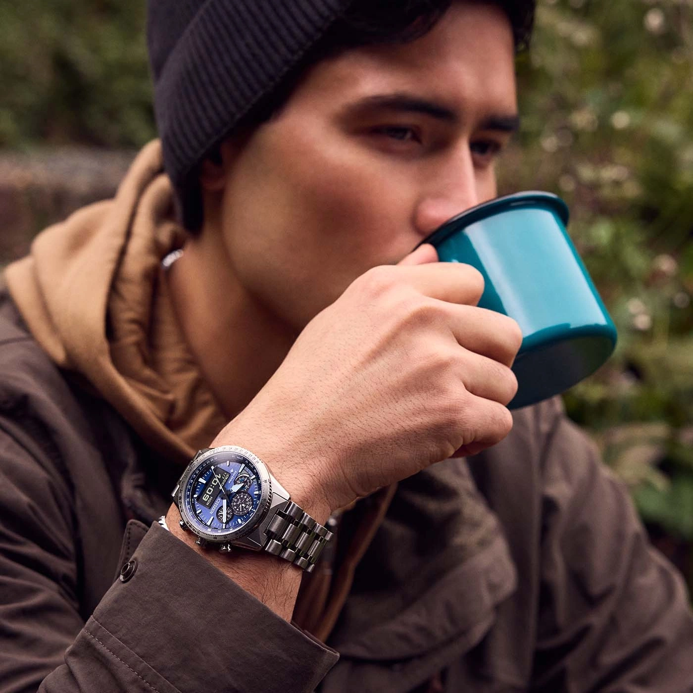 Man drinkt uit een mok, draagt een horloge en een muts in de natuur.