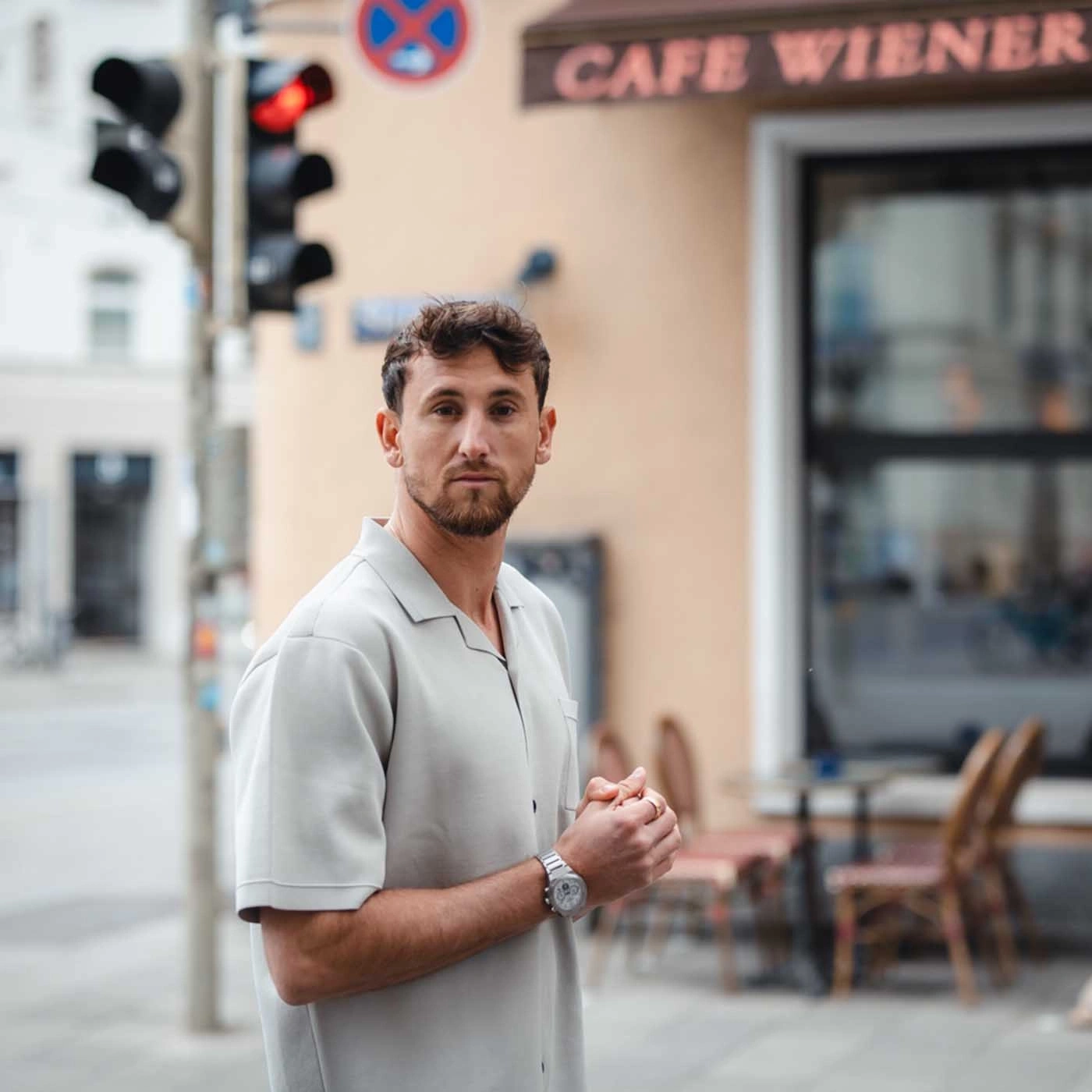 Man voor een café, draagt een licht overhemd en een horloge, kijkt naar de camera.