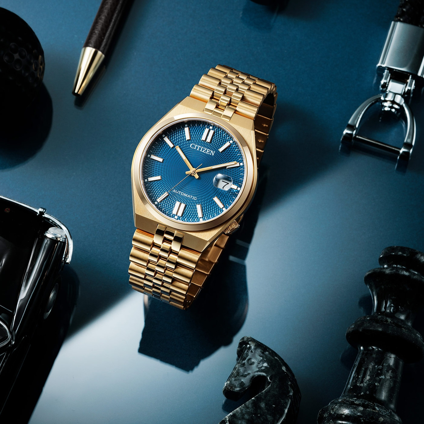 Gouden Citizen horloge met blauwe wijzerplaat, pen, schaakstukken en sleutelhanger op blauw oppervlak.