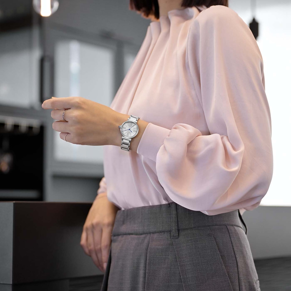 Vrouw in roze blouse en grijze broek kijkt op haar horloge.