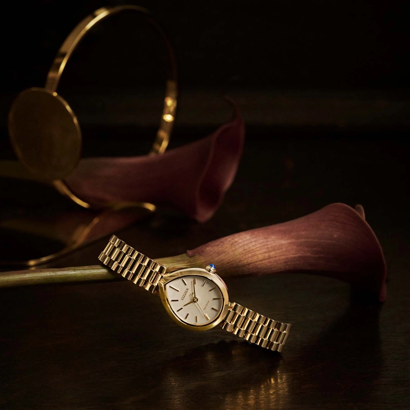 Een gouden horloge met band rust op donker oppervlak met bloemen.