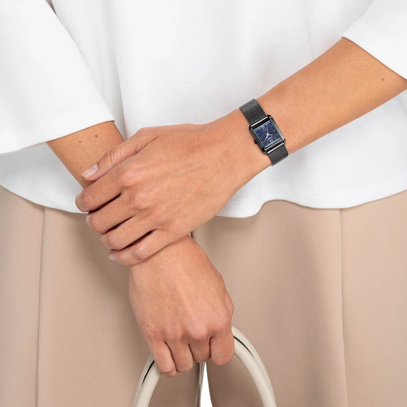 Vrouw met zwart horloge, witte tas, beige broek, wit overhemd.