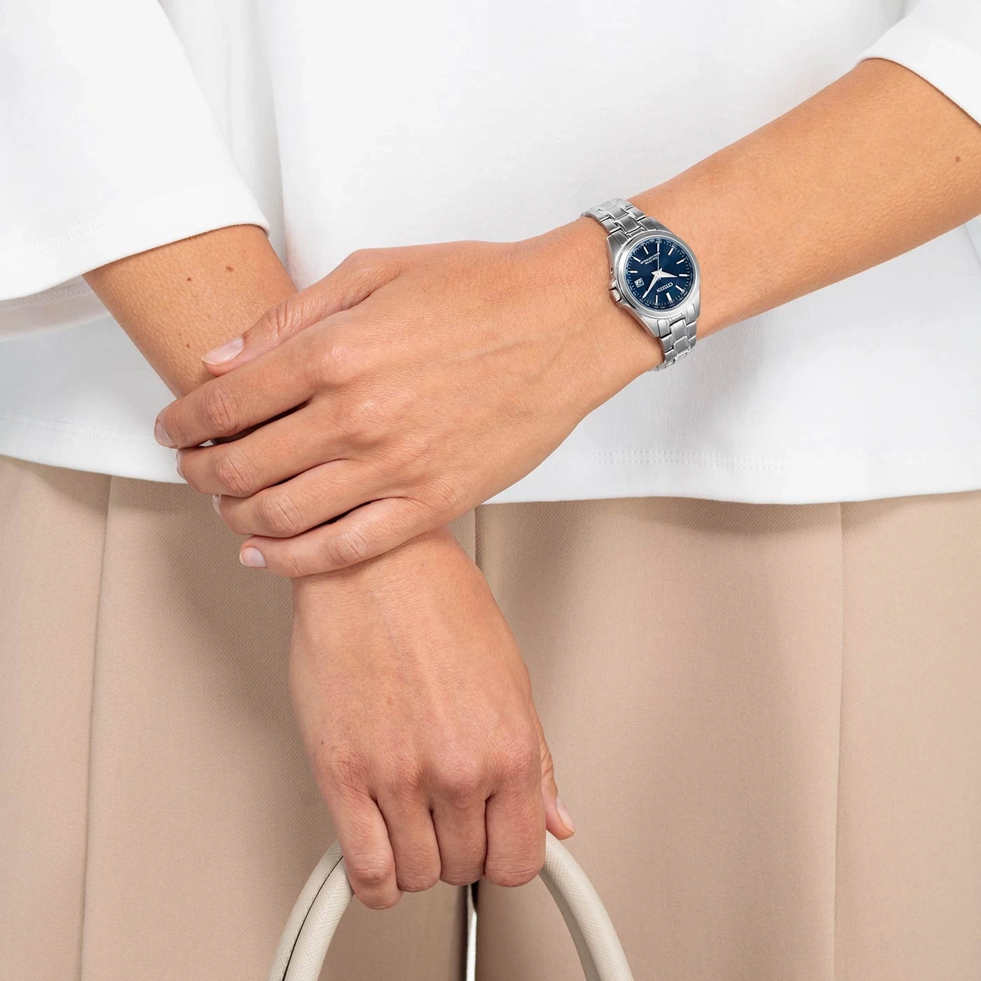 Vrouw met horloge, tas, wit shirt en beige broek.