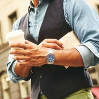 Man met koffie en krant, draagt horloge en vest, buiten.