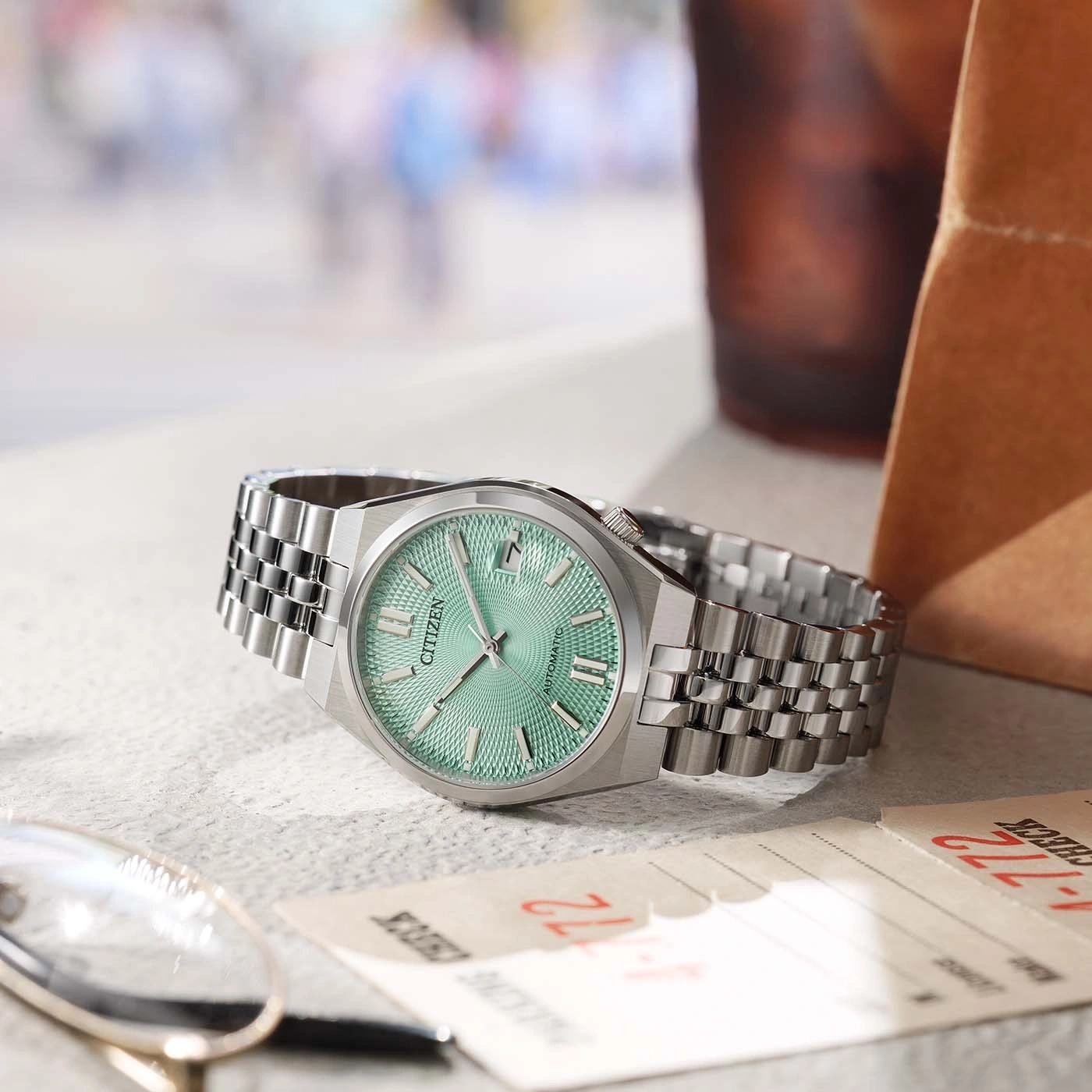 Een Citizen automatisch horloge met een mintgroene wijzerplaat rust op een getextureerd oppervlak naast een bril en papieren.