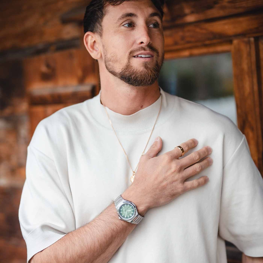 Man met horloge, ring en ketting, raakt zijn borst aan, kijkt weg, houten achtergrond.