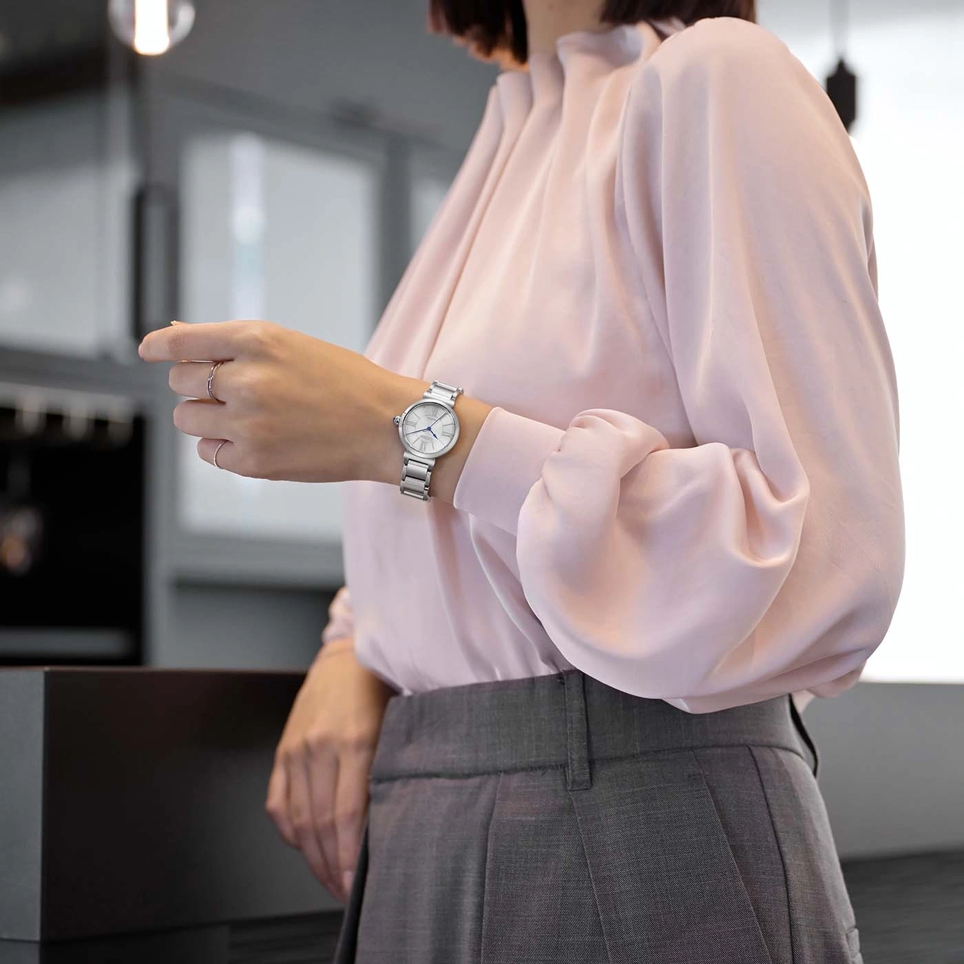 Vrouw in roze blouse en grijze broek kijkt op haar horloge.