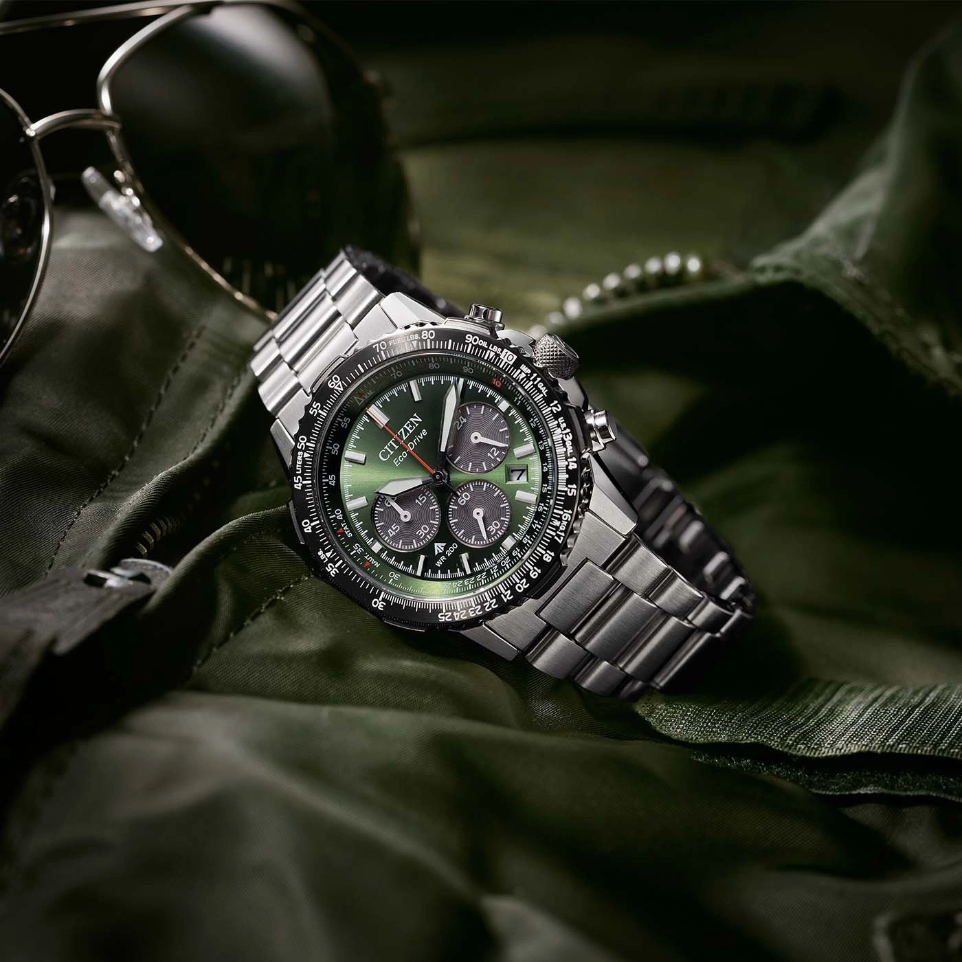 Citizen horloge met groene wijzerplaat en metalen band op groene stof met zonnebril.