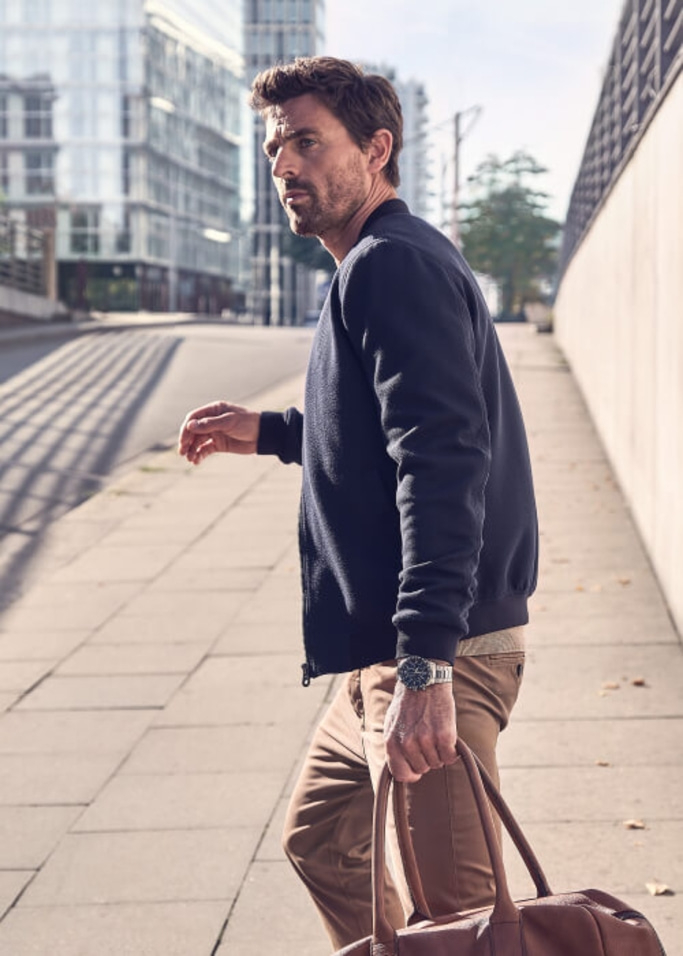 Man loopt op de stoep, houdt een tas vast, draagt een jas en een horloge.