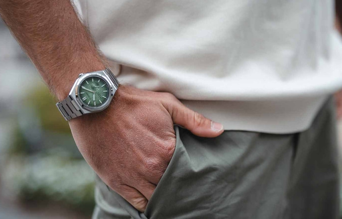Man met een horloge met groene wijzerplaat, hand in zak, casual stijl.