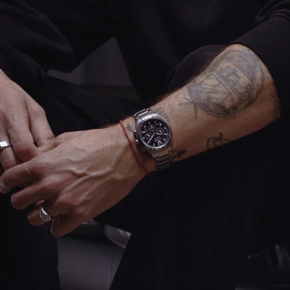 Close-up van een getatoeëerde arm van een man met een horloge en ringen.