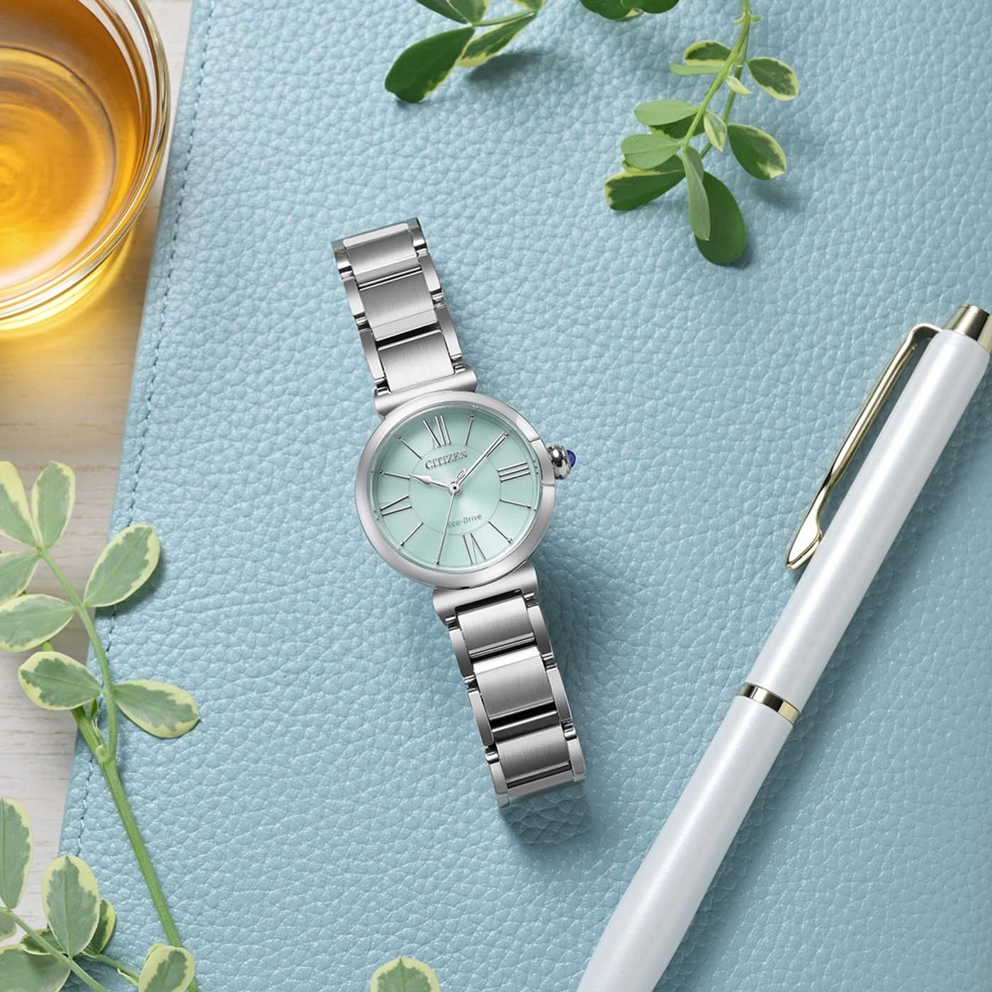Een mintgroen Citizen Eco-Drive horloge met zilveren band ligt op een blauw getextureerd oppervlak naast een witte pen en een glas amberkleurige vloeistof.