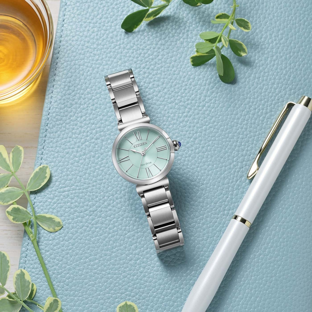 Een mintgroen Citizen Eco-Drive horloge met zilveren band ligt op een blauw getextureerd oppervlak naast een witte pen en een glas amberkleurige vloeistof.