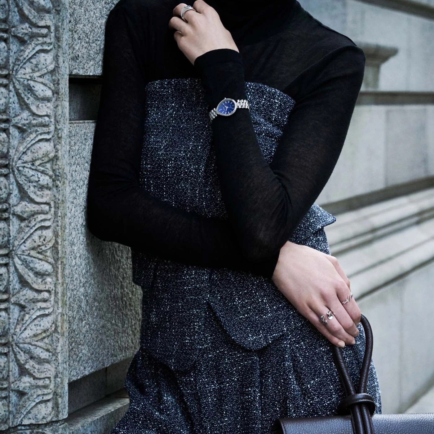 Vrouw in zwarte coltrui en jurk met textuur, draagt een horloge en ringen.