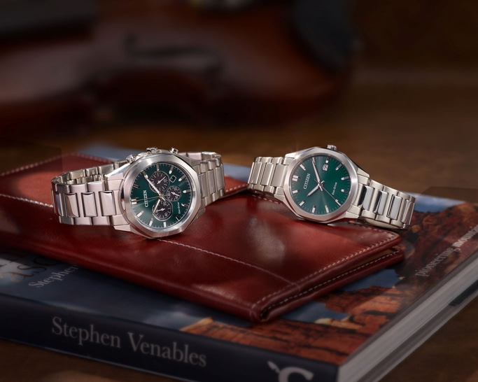 Twee Citizen horloges met groene wijzerplaten op een boek en leren portemonnee.