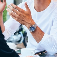 Man met horloge, gebarend met handen, zittend aan een tafel met koffiekopjes.