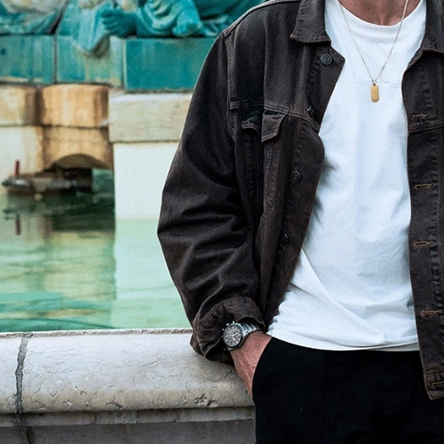 Man in bruine jas en wit shirt, horloge, leunend op een muur