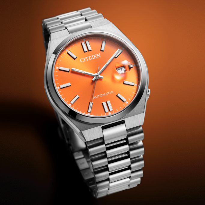 A stylish Citizen automatic watch with a vibrant orange dial and stainless steel bracelet, presented against a warm, gradient background.
