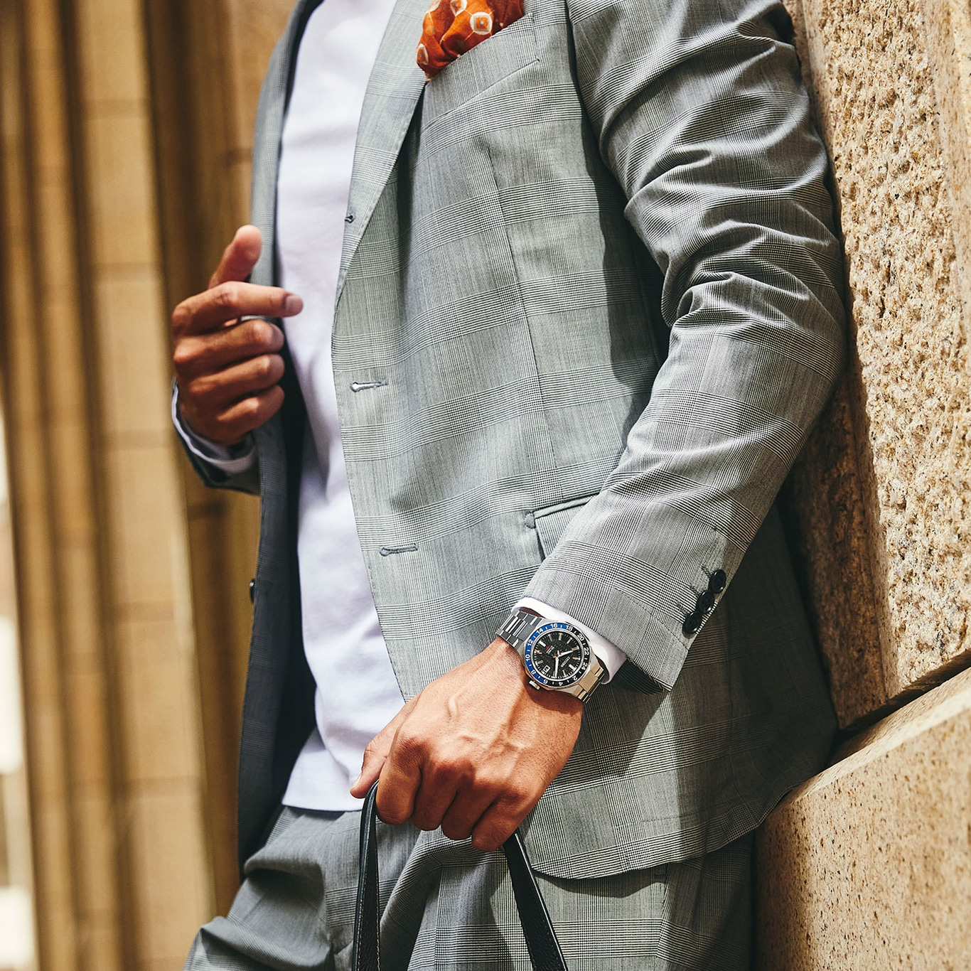Man in pak met horloge en pochet leunt tegen een muur