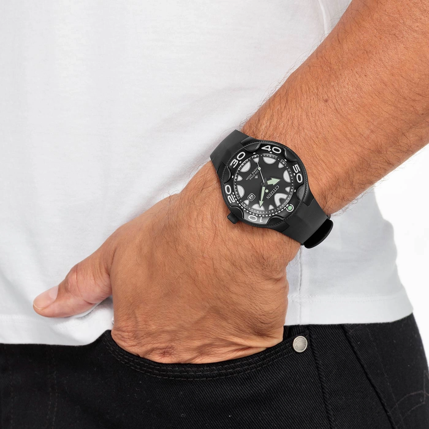 Man met zwart horloge, wit shirt en zwarte jeans, hand in de zak.