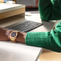 Gouden horloge om de pols, groene trui, toetsenbord, witte bureau, kantooromgeving.