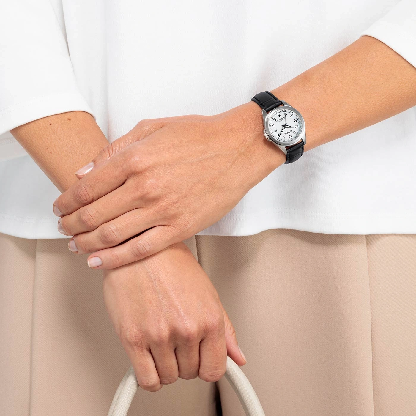 Vrouw met horloge, houdt een tas vast, met wit overhemd en beige broek.