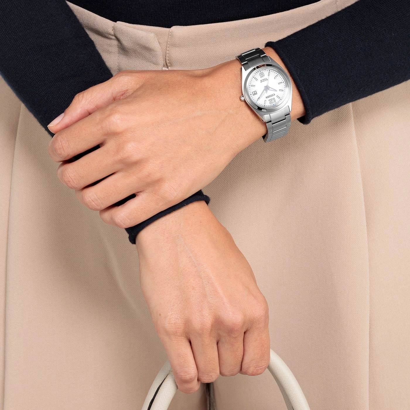 Vrouw met zilveren horloge, houdt een witte handtas vast, beige broek.