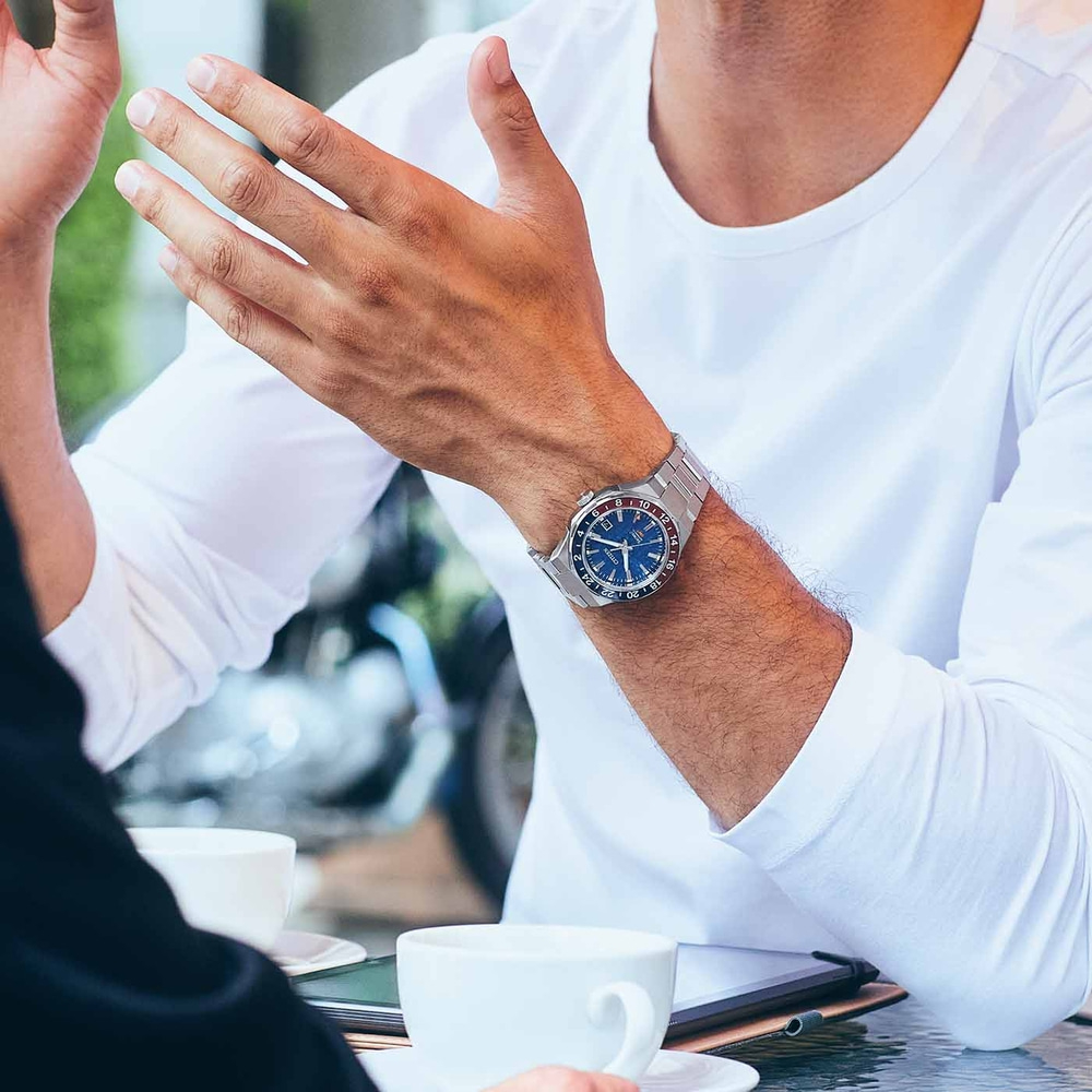 Man met horloge, gebarend met handen, zittend aan een tafel met koffiekopjes.