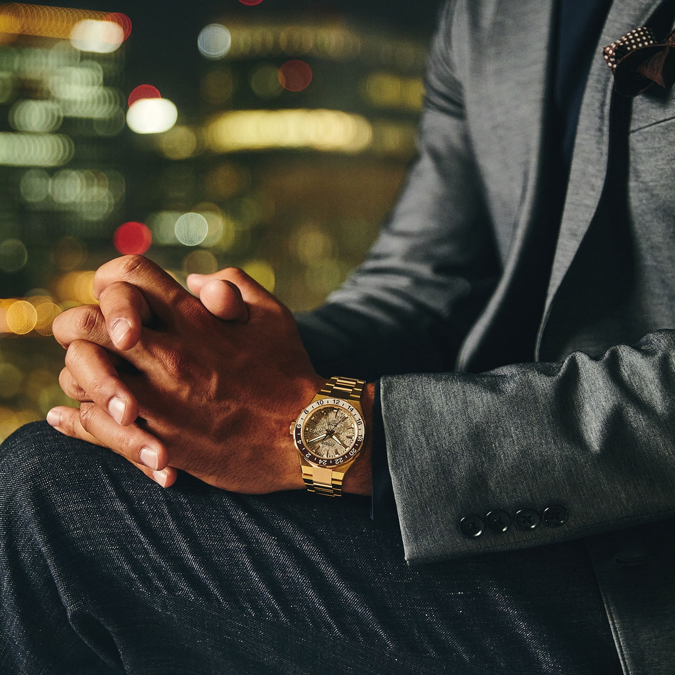 Man in pak met gouden horloge, handen gevouwen, stadslichten op de achtergrond.