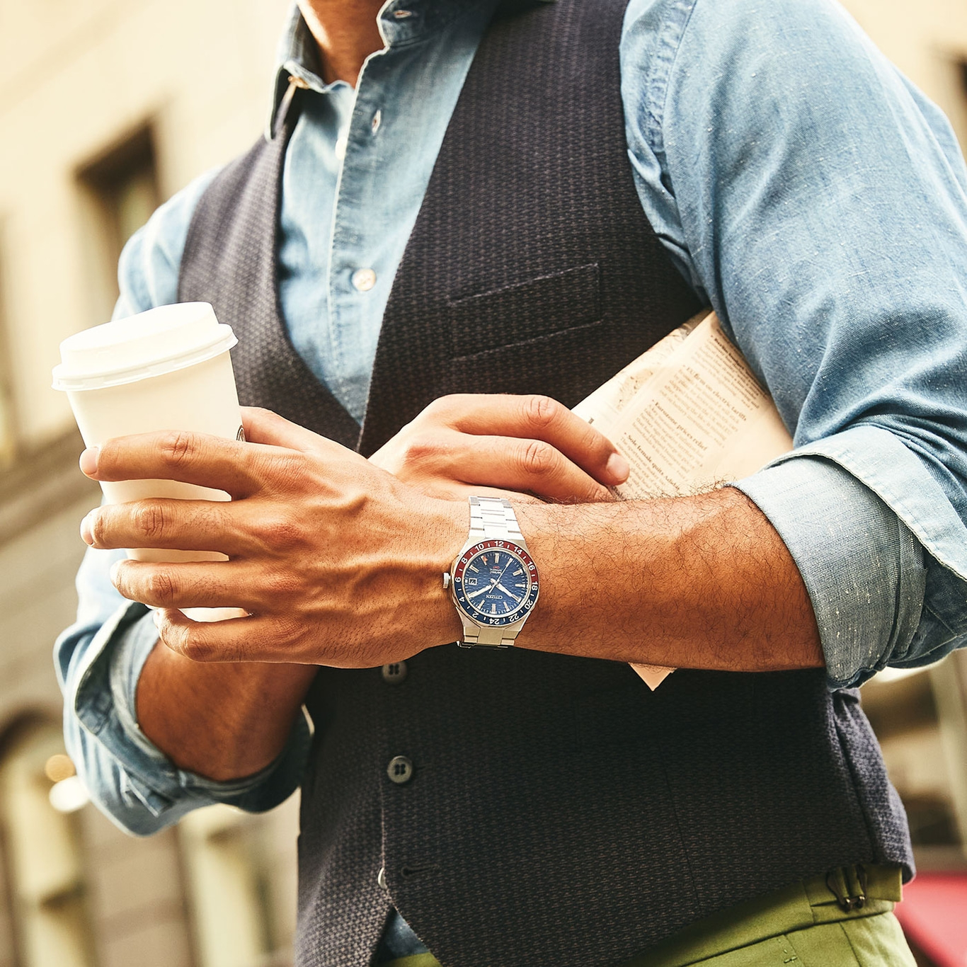 Man met koffie en krant, draagt horloge en vest, stedelijke omgeving.
