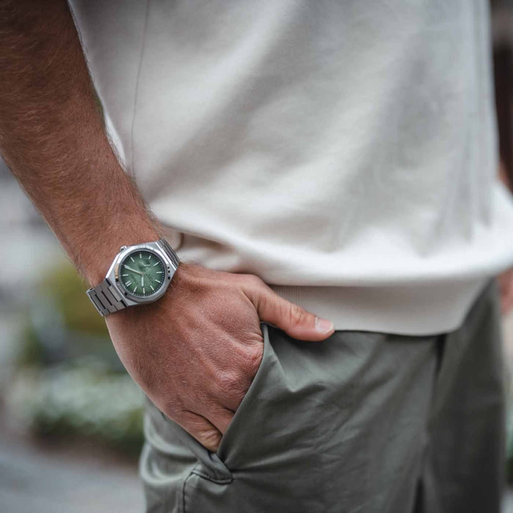 Man draagt zilveren horloge met groene wijzerplaat, hand in zak.