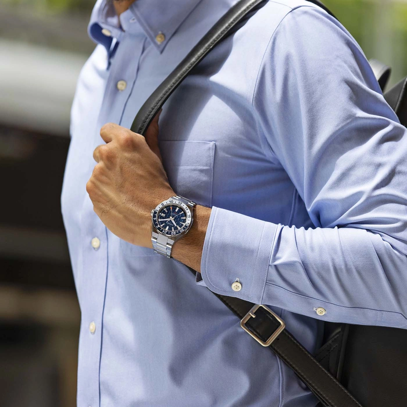 Man met horloge en blauw overhemd met een zwarte tas op zijn schouder.