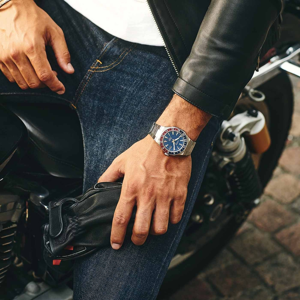 Man met horloge, leren jas en jeans, houdt handschoenen vast op een motorfiets.