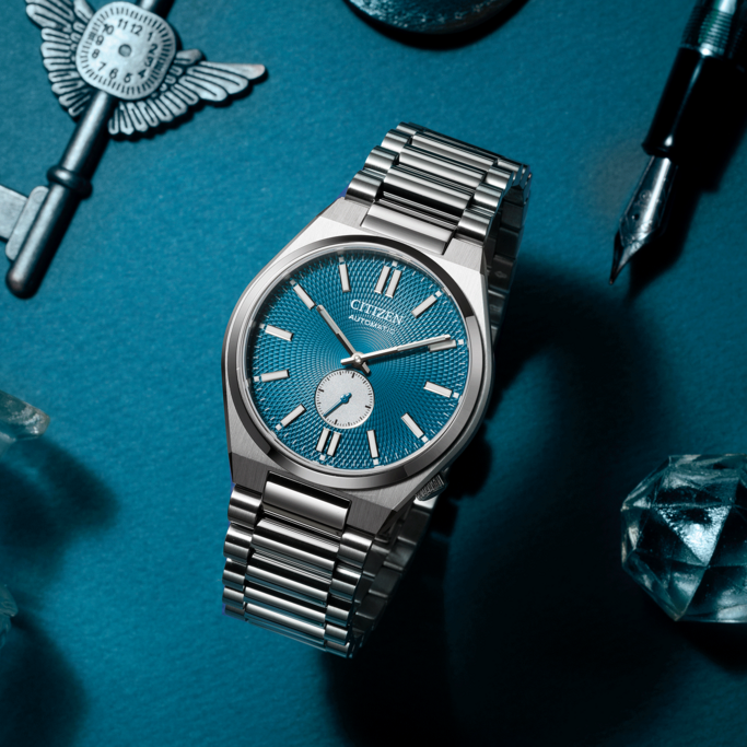 Citizen horloge met blauwe wijzerplaat, sleutel, pen en kristallen op blauw oppervlak.